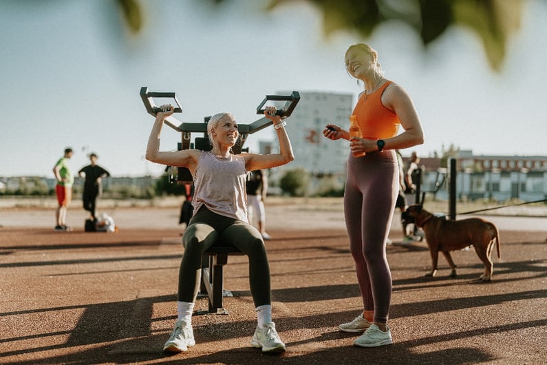 Två kvinnor tränar på ett utegym; en kvinna använder en träningsmaskin medan den andra håller en vattenflaska och skrattar.