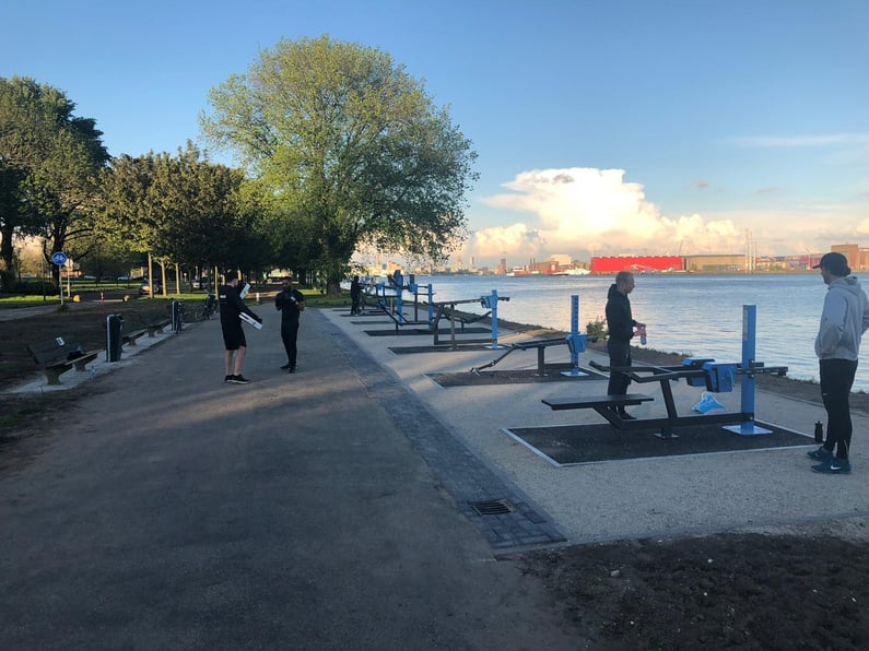 Utegym vid vattnet med människor som tränar och promenerar, omgiven av träd och med en stadssilhuett i bakgrunden.